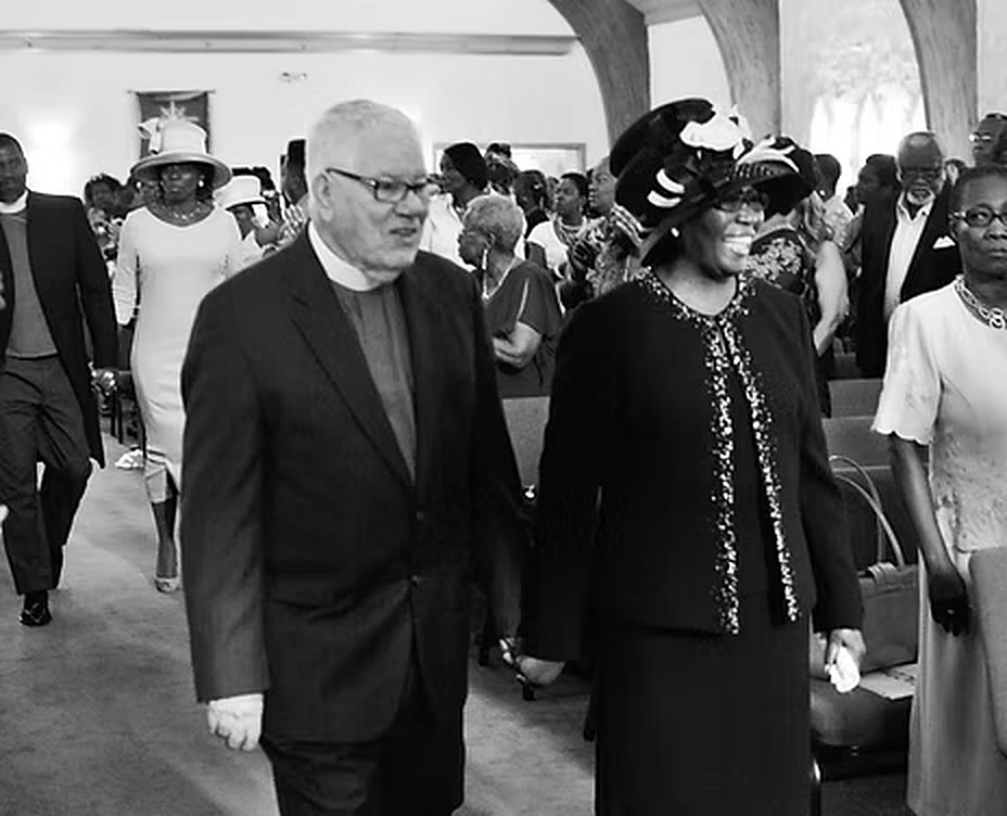 Overseer Stanley Kidd and Rev. Lenore Kidd walk the center aisle together as the congregation stands, in black and white.